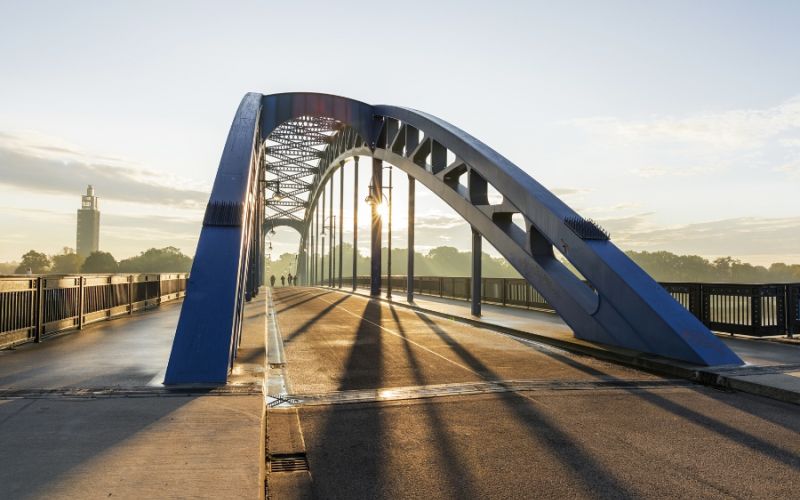 Blick auf Jerusalembrücke in Magdeburg bei Sonnenaufgang mit markanten blauen Bogendesign. Sie verfügt über einen Fußgängerweg mit Geländern auf beiden Seiten.